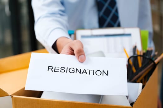 Close-Up of Man's Hand Delivering Resignation Letter with Box of Personal Belongings in the Background, Indicating Professional Departure