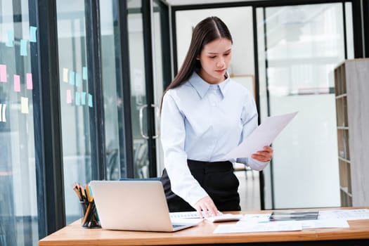 A focused female professional intently reviewing a document at her office desk with a laptop and smartphone nearby.