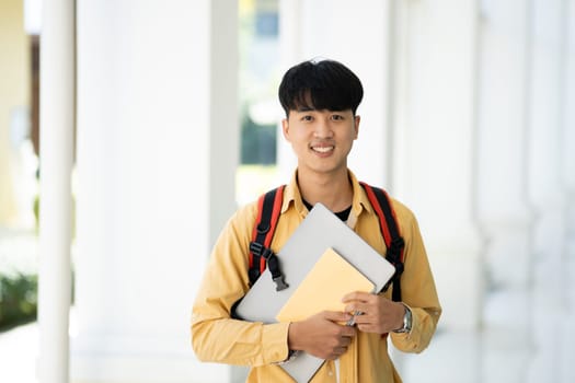 A college student stands in the hallway of his school, holding textbooks and smiling, ready for a day of learning and studies.