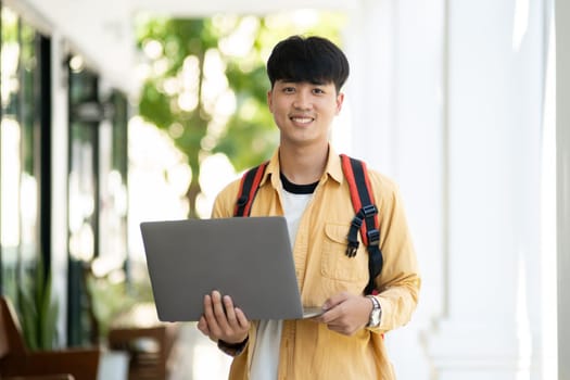 A male student with a pleasant smile stands in the school hallway, confidently holding his laptop, ready for class.