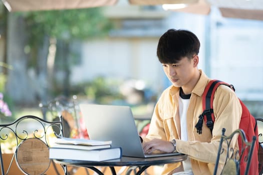 A university student is absorbed in thought while studying on his laptop, sitting at an outdoor table surrounded by the tranquility of the campus.