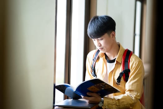 A young man is reading a book while wearing a yellow shirt and a red backpack. He is sitting by a window, and the book he is reading is blue