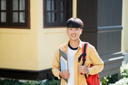 A happy and content young man stands outside on his university campus, holding a laptop and books, embodying the optimism of college life.