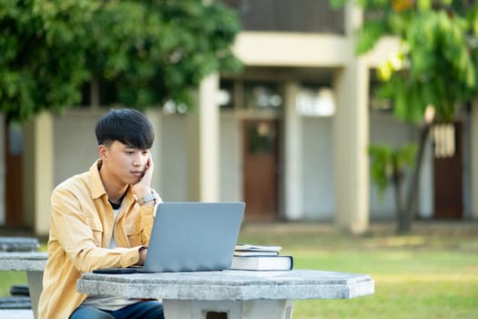 A university student is absorbed in thought while studying on his laptop, sitting at an outdoor table surrounded by the tranquility of the campus.