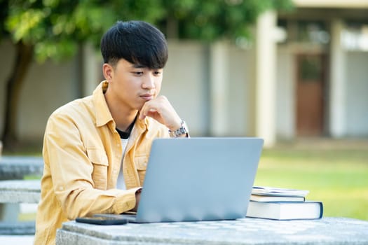 A university student is absorbed in thought while studying on his laptop, sitting at an outdoor table surrounded by the tranquility of the campus.