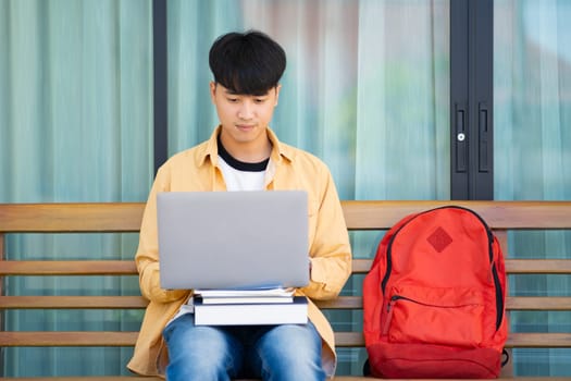 A college student sits on an outdoor bench, working intently on his laptop, with a stack of books and a red backpack by his side.