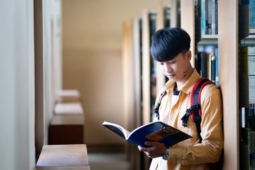 A young man is reading a book in a library. He is wearing a backpack and has a serious expression on his face. The library is quiet and peaceful