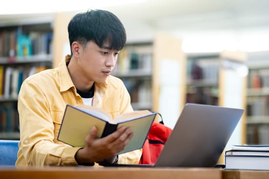A young man is sitting at a desk with a laptop and a book. He is reading the book while looking at the laptop. The scene suggests that he is studying or working on a project