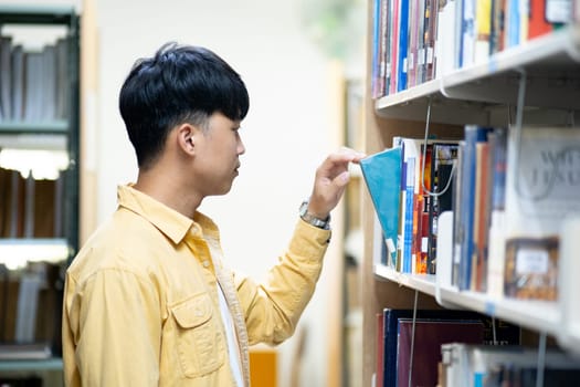A man in a yellow shirt is looking at a book on a library shelf. He is reaching for a book with a blue cover