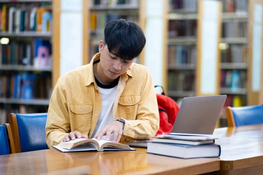A young man is sitting at a table in a library, reading a book. He is wearing a yellow shirt and a black shirt. There are several books on the table, including a laptop