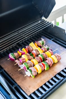 Skewered beef and fresh veggies sizzle on a copper grill mat over a gas outdoor grill.