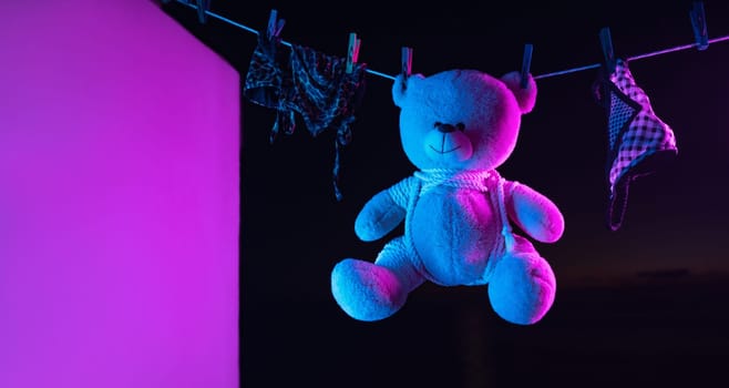 toy teddy bear in a bdsm mask and leather straps hangs on a clothesline in neon light on a dark background