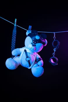 toy teddy bear in a bdsm mask and leather straps hangs on a clothesline in neon light on a dark background