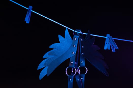 bdsm leather shoulder straps in the shape of wings hanging on clothesline in neon light on a black background, conceptual photos