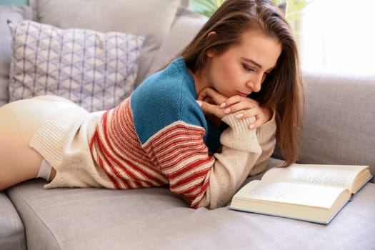 Young model, reading a book on the couch, lying low, dressed in a sweater.