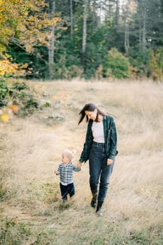 Mom leads a little girl by the hand across the lawn, looking at her with a smile. High quality photo