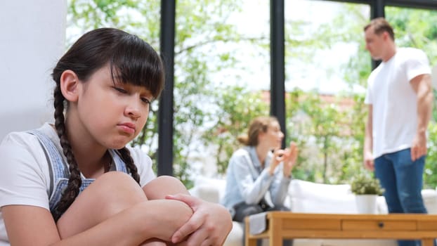 Stressed and unhappy young girl huddle in corner, cover her ears blocking sound of her parent arguing in background. Domestic violence at home and traumatic childhood develop to depression. Synchronos