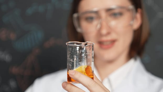 Closeup of young beautiful teacher holding chemical liquid at laboratory. Scientist doing an experiment by inspect colored solution while standing in front of blackboard. Focus on beaker. Erudition.