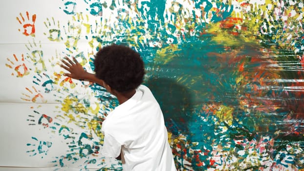 Back view of happy african boy painted the stained wall with colorful hand while wearing casual white shirt in art lesson.Smart student use hand print to make creative artwork. Education. Edification.