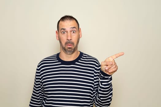 Hispanic man with beard in his 40s wearing a striped sweater pointing to the side with finger with a surprised face, isolated on beige background