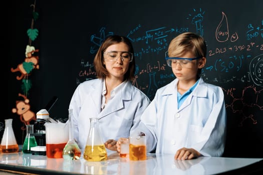 Skilled teacher watch out schoolboy in laboratory wear lab coat and glasses stand and experiment about science of chemistry in STEM class. Teacher pour red liquid down to yellow beaker. Erudition.