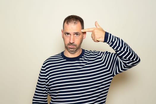 Hispanic man with beard in his 40s wearing a striped sweater standing over a beige background shooting and killing himself pointing with hand and fingers at his head like a gun, suicidal gesture.