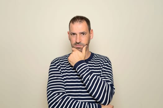 Hispanic man with beard about 40 years old wearing striped sweater thinking concentrated in doubt with finger on chin and looking at the camera wondering, isolated over beige background