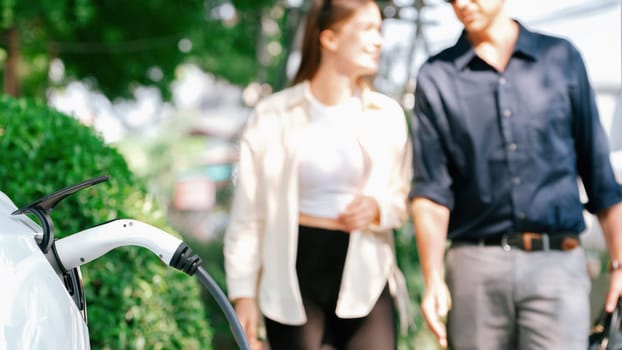 Young couple recharge electric car battery from charging station in green city park in springtime. Rechargeable EV car for sustainable environmental friendly urban travel lifestyle. Panorama Expedient