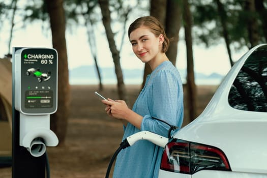 Holiday road trip vacation traveling to the beach camp with electric car, young woman checking battery from smartphone while recharge EV vehicle. Beach travel camping with eco-friendly car .Perpetual