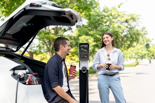 Lovely young couple drinking coffee while recharging battery for electric car during road trip travel EV car in natural forest or national park. Eco friendly travel during vacation and holiday. Exalt