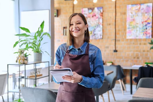 Successful young woman service worker owner in apron with working notepad pen looking at camera in restaurant cafeteria coffee pastry shop interior. Small business staff occupation entrepreneur work