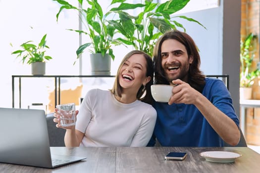 Happy joyful laughing young couple looking at laptop together while sitting in cafeteria. Leisure time for two, communication, togetherness, relationship, lifestyle, work, study remotely