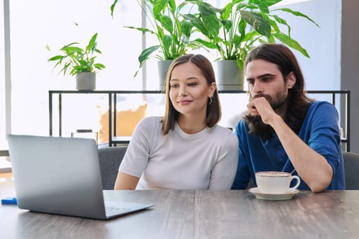 Calm serious young couple looking at laptop together while sitting in cafe, cafeteria. Leisure time for two, lifestyle, togetherness, relationship, communication, work study remotely