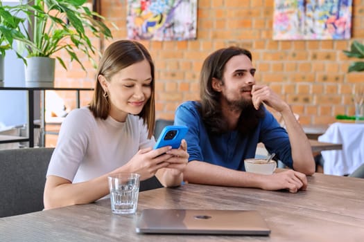 Young handsome people friends, couple man and woman relaxing together in a cafe, using smartphone, drinking coffee. Lifestyle, leisure, youth concept