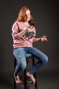 A redhead youn woman is sitting in a stool, holding her smartphone, while she smiles and looks for new contents and messages.