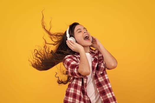 Amazed teenager. Teenager child girl in headphones listening music, wearing stylish casual outfit isolated over yellow background. Excited teen girl