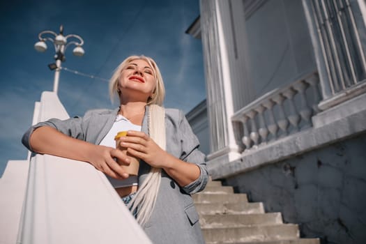 A blonde woman in a suit is holding a coffee cup and looking up at the sky. Concept of sophistication and elegance, as the woman is dressed in a business suit and holding a coffee cup