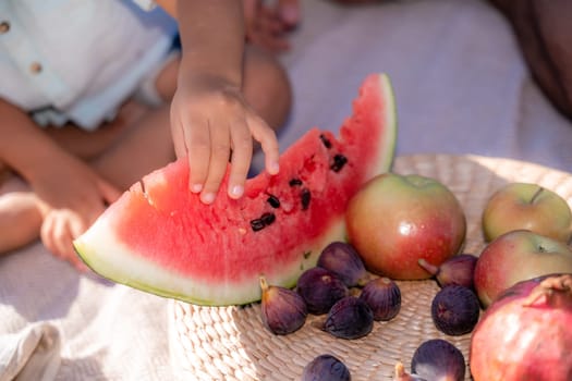 A child is eating a slice of watermelon while sitting on a blanket. There are also apples and figs on the blanket