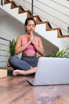 Vertical portrait of young Asian woman doing online meditation with hands in prayer at home living room sitting on yoga mat using laptop. Spirituality concept.