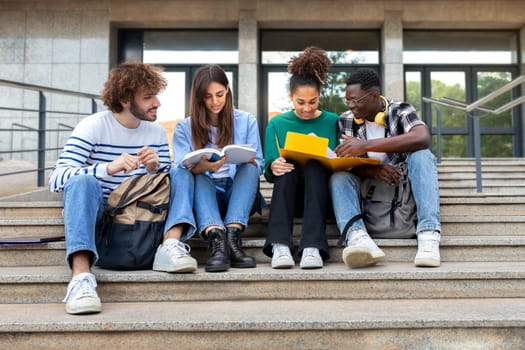 Happy diverse university student friends doing homework together sitting on campus steps. Higher education and technology concept.