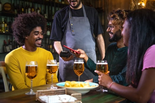 Male customer paying beer round in a bar with mobile phone. Friends paying drinks with phone at pub. Technology and leisure concept.