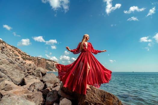 Red dress sea woman. A blonde with flowing hair in a long flowing red dress stands on a rock near the sea. Travel concept, photo session at sea.