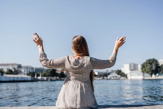 A woman in a white dress is sitting on a ledge by a body of water, with her arms outstretched. Concept of freedom and relaxation, as the woman is enjoying the peaceful surroundings