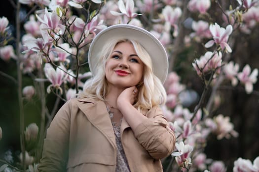 Magnolia flowers woman. A blonde woman wearing a hat stands in front of a tree with pink magnolia flowers. She is wearing a tan coat and a dress