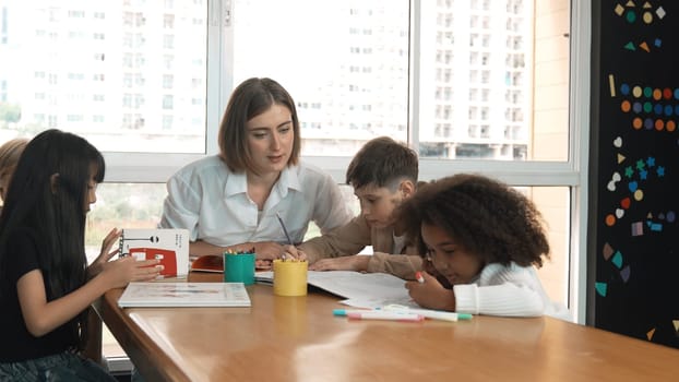 Professional caucasian teacher telling story to diverse student while sitting at table with storybook and colored book. Smart learner listening story while colored picture from instructor. Erudition.