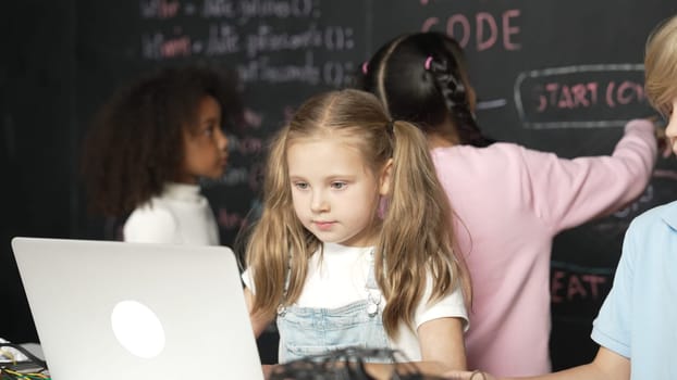 Smart girl using laptop programing engineering code with diverse friends ay table with microchip and main board scatter around in STEM classroom at blackboard written with coding prompt. Erudition.