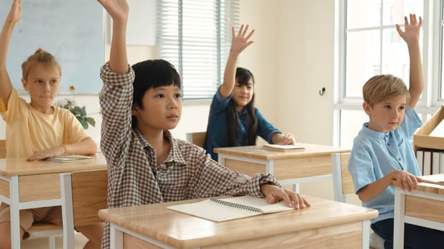 Smart boy raising hand for asking and answering teacher surrounded by multicultural children studying at classroom. Happy student working together, voting, volunteering, calling instructor. Pedagogy.