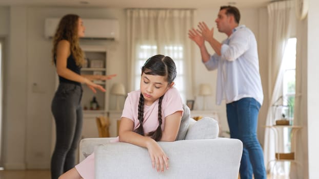 Annoyed and unhappy young girl sitting on sofa trapped in middle of tension by her parent argument in living room. Unhealthy domestic lifestyle and traumatic childhood develop to depression Synchronos