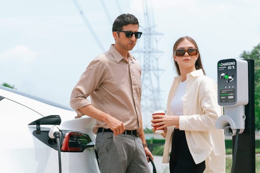 Young couple with coffee recharge EV car battery at charging station connected to electrical industrial power grid tower. Couple with shopping and travel using eco electric car lifestyle. Expedient