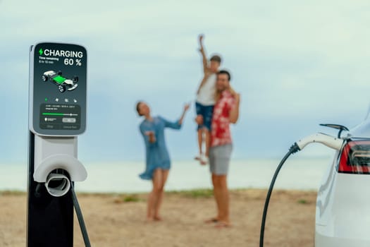 Alternative family vacation trip traveling by the beach with electric car recharging battery from EV charging station with blurred cheerful and happy family enjoying the seascape background. Perpetual